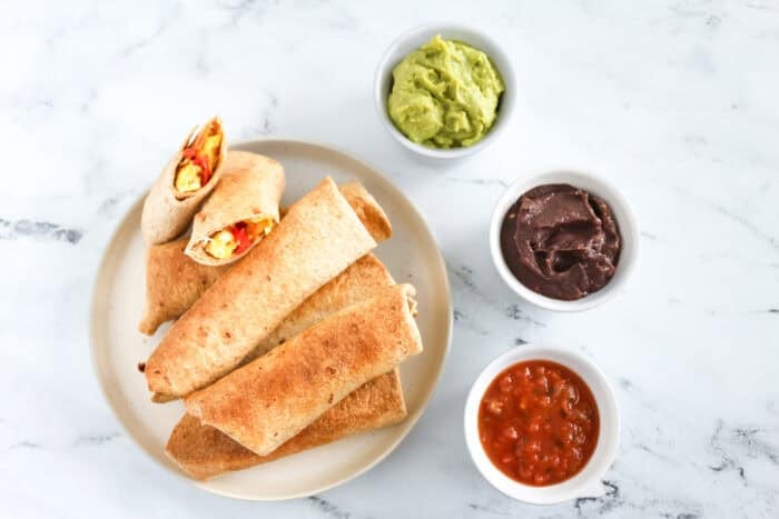 Plate of baked breakfast taquitos filled with scrambled eggs and peppers, served with &iexcl;Yo Quiero! Grab & Go Guacamole, Black Bean Dip, and Salsa.