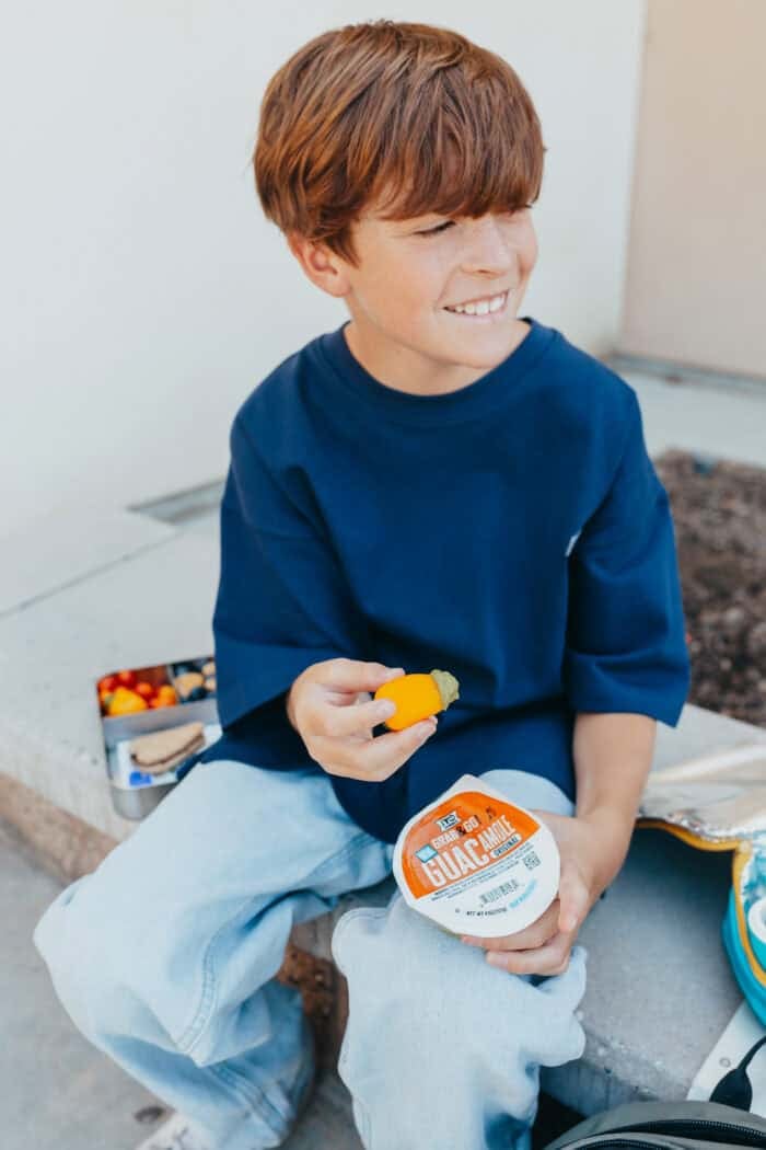 Smiling boy sitting outdoors holding a yellow pepper dipped in guacamole, with a &iexcl;Yo Quiero! Grab & Go Guacamole cup in hand.