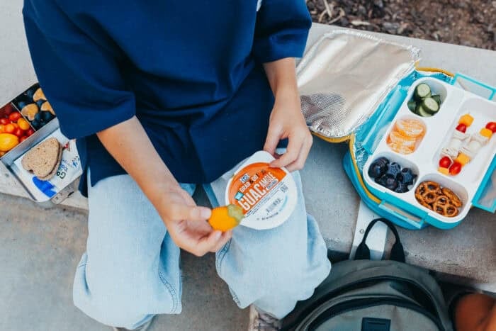 A boy sitting outdoors holding a yellow pepper dipped in guacamole, with a &iexcl;Yo Quiero! Grab & Go Guacamole cup in hand.