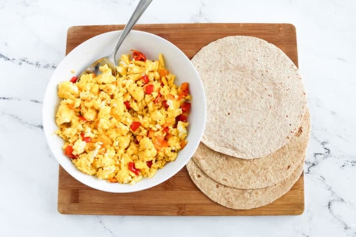 Bowl of scrambled eggs with chopped bell peppers next to a stack of whole wheat tortillas on a wooden cutting board.