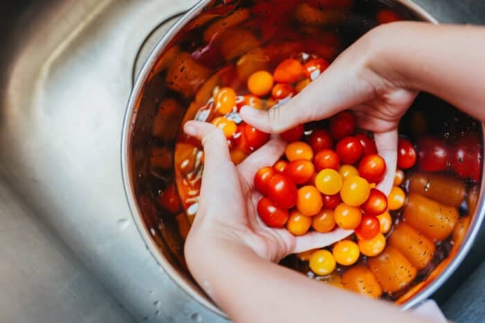 Close-up of hands rinsing Sunset® Sprinkles teeny tiny tomatoes in a large bowl of water over a kitchen sink.
