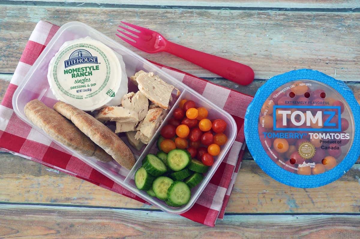 Bento box filled with chicken, pita bread, small tomatoes, cucumber slices and Ranch dip, placed on red checkered napkin.