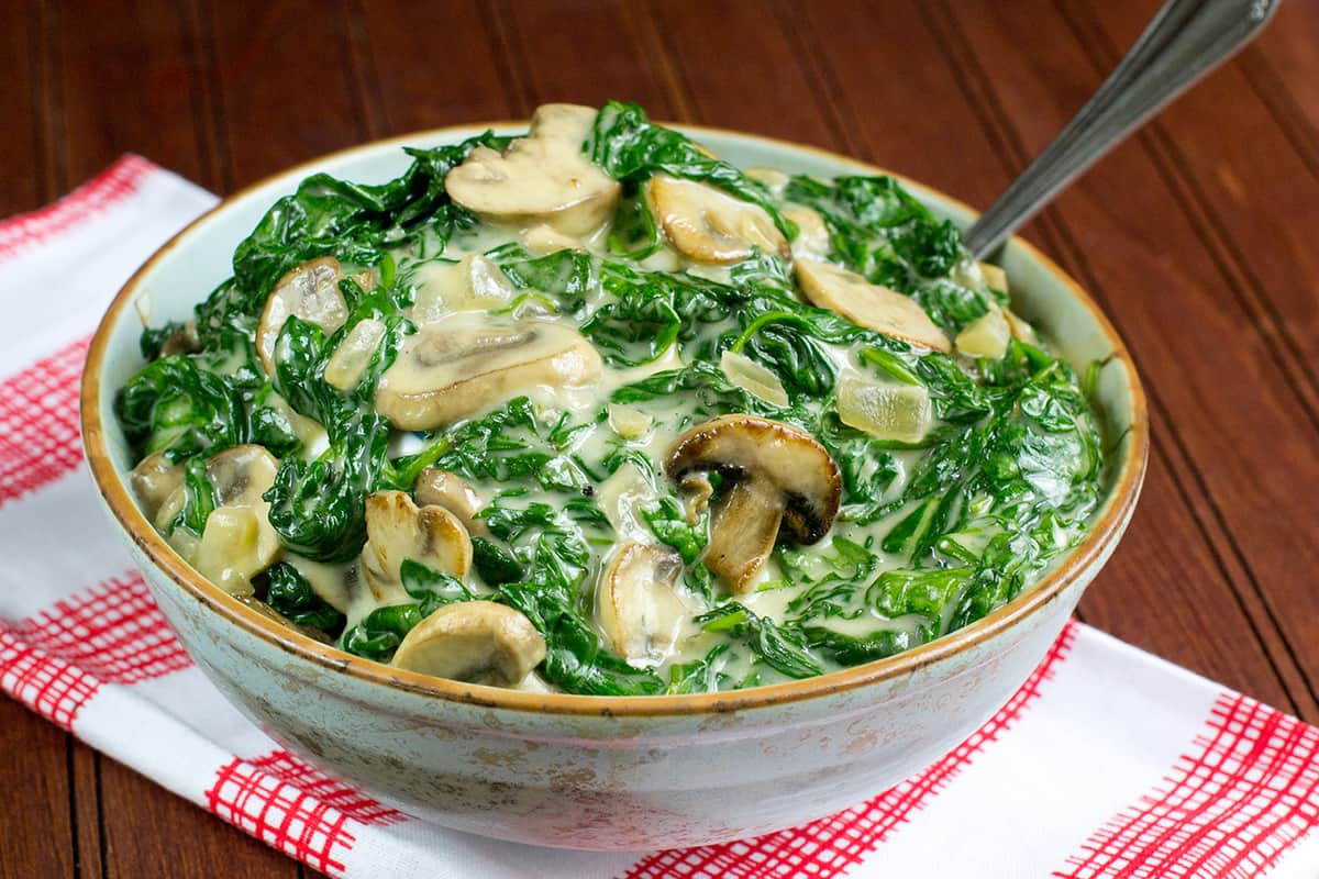 Bowl of creamed spinach with mushrooms on red and white napkin placed on wooden table.